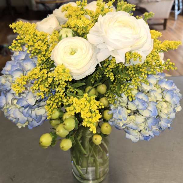 Bouquet of white ranunculus, blue hydrangea, and yellow filler in a glass vase