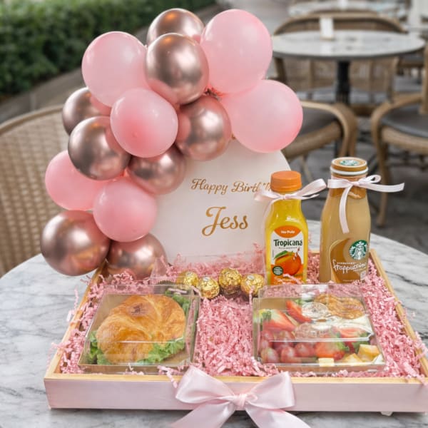 Birthday gift tray with pink and rose-gold balloons, snacks, and drinks