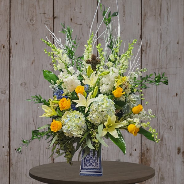 Tall floral arrangement with white lilies, yellow roses, and blue hydrangeas in a patterned vase