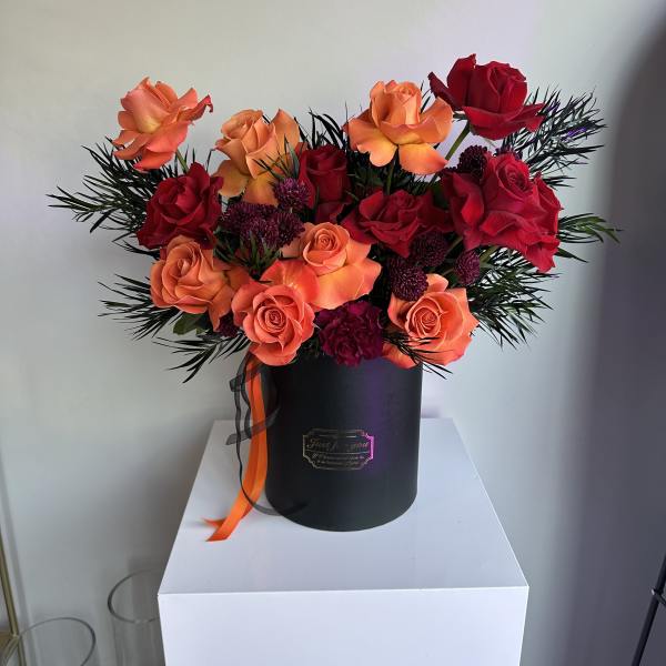 Orange and red roses with dark accents arranged in a black round hatbox with ribbon