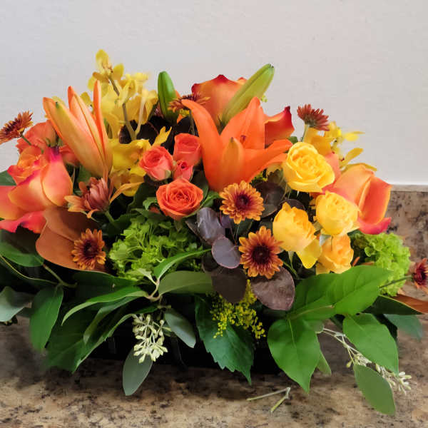 Orange and yellow floral arrangement with roses and lilies in a low container