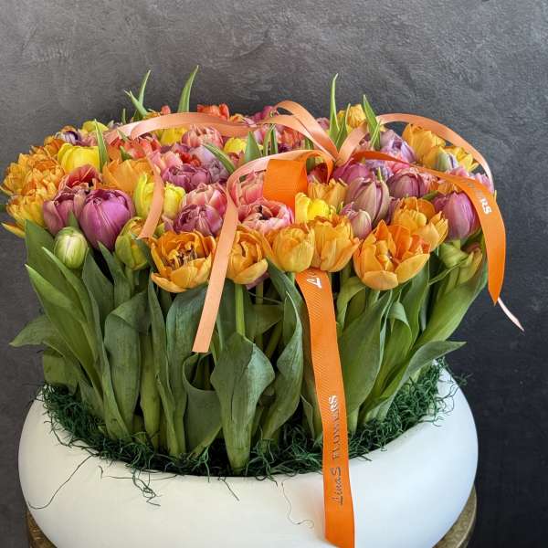 Low white bowl packed with orange, yellow, and pink tulips topped with orange ribbons