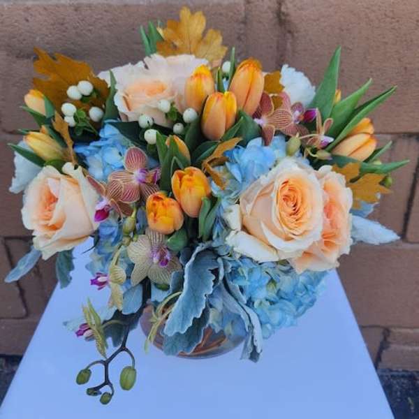 Low arrangement of peach roses, orange tulips, blue hydrangeas, and orchids in a clear glass vase