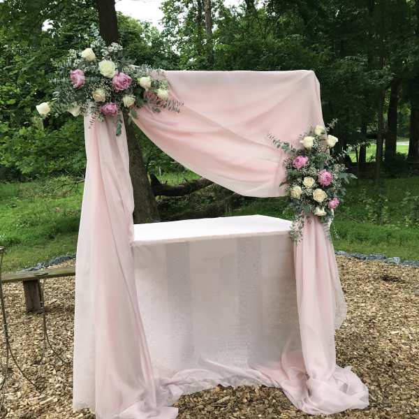 Pink draped wedding arch with floral arrangements and a table beneath it
