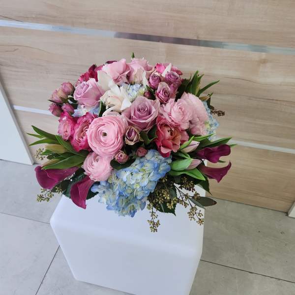 Rounded bouquet of pink roses, ranunculus, lilies and blue hydrangeas in a compact arrangement.