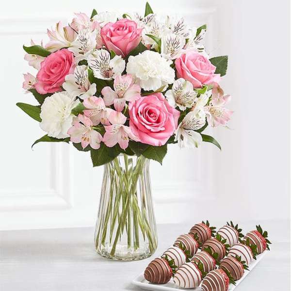 Pink and white bouquet in a clear glass vase with chocolate-covered strawberries on a tray