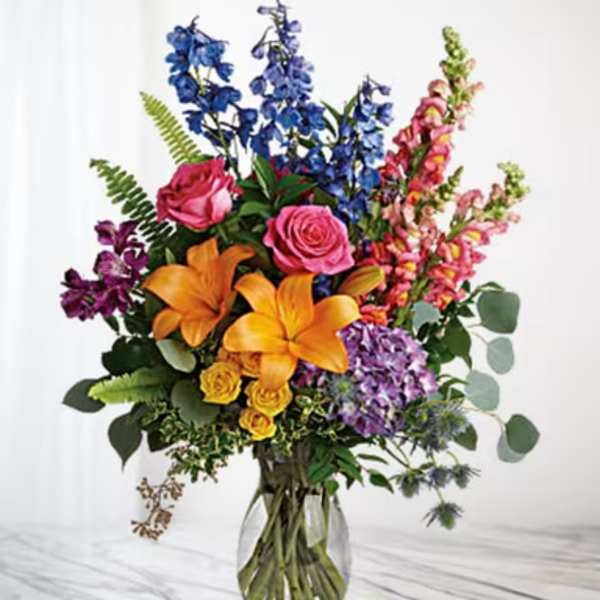 Colorful bouquet with lilies, roses, and tall blue and pink blooms in a glass vase