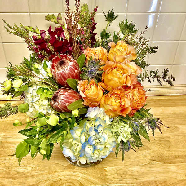 Mixed bouquet with orange roses, blue hydrangea, and burgundy protea in a low vase