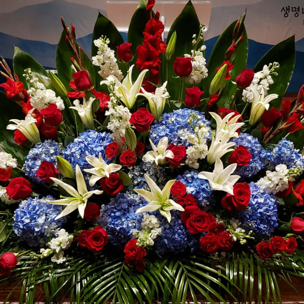 Large red, white, and blue floral spray with roses, lilies, hydrangeas, and bold green leaves.