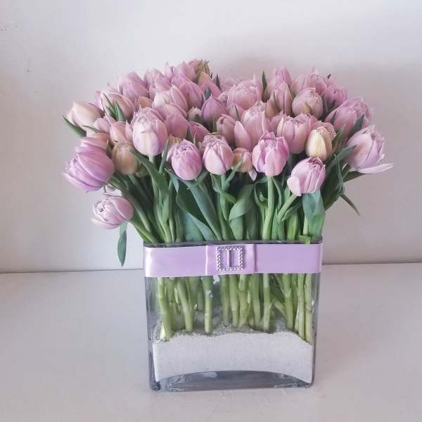 Pink tulips arranged in a clear square vase with a lavender ribbon