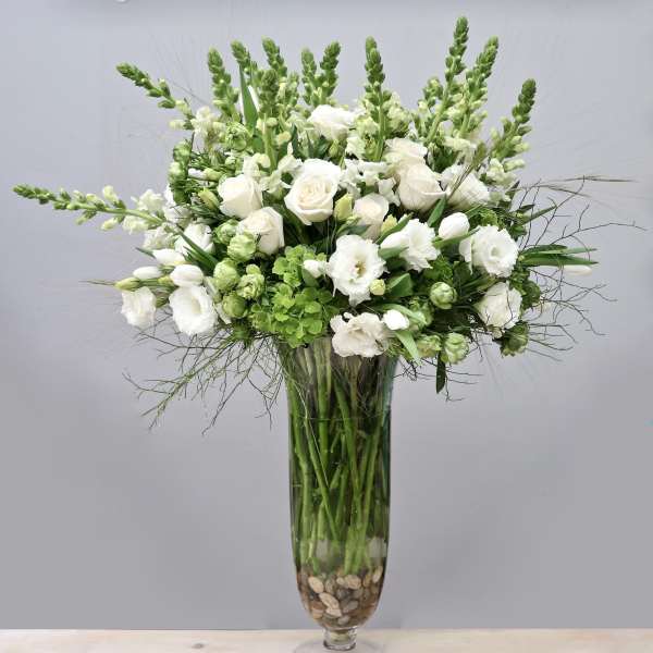 Tall white and green floral arrangement with roses and snapdragons in a clear glass vase with stones