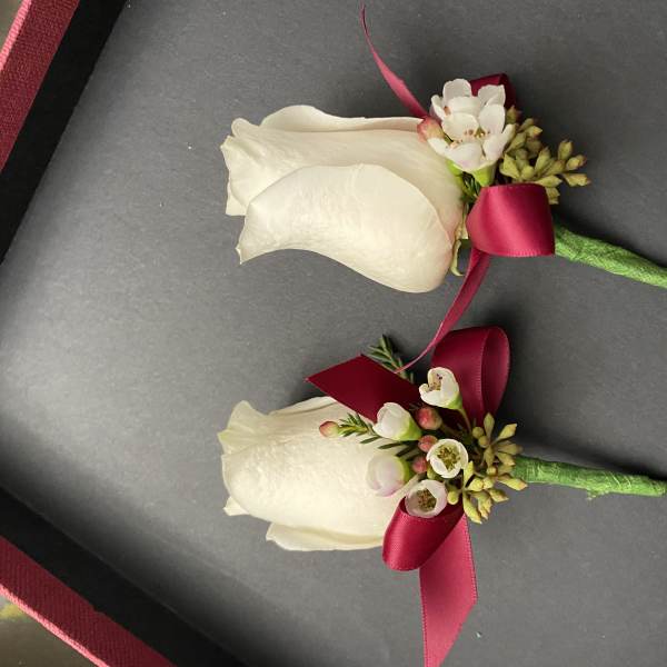 Two white rose boutonnieres with burgundy ribbon accents