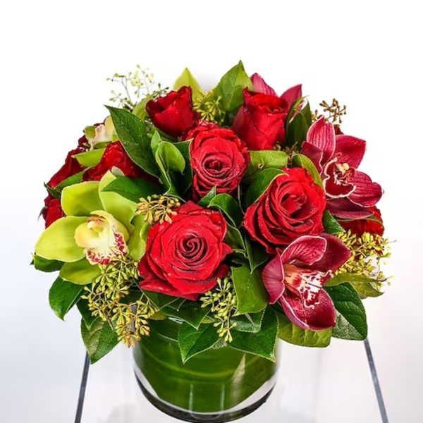 Round arrangement of red roses and orchids in a clear glass cylinder vase wrapped with green leaves