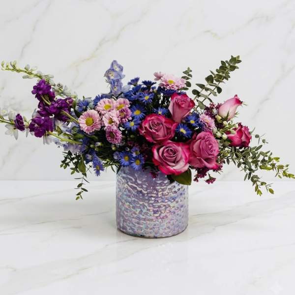 Asymmetrical arrangement of pink roses and purple and blue blossoms in an iridescent lavender vase