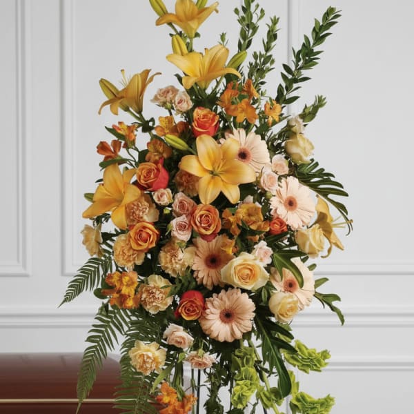 Tall standing spray with yellow lilies and peach roses on an easel beside a wooden casket.