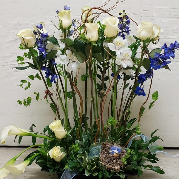 Tall arrangement of white roses, calla lilies, and blue flowers with a small bird nest in a low tray