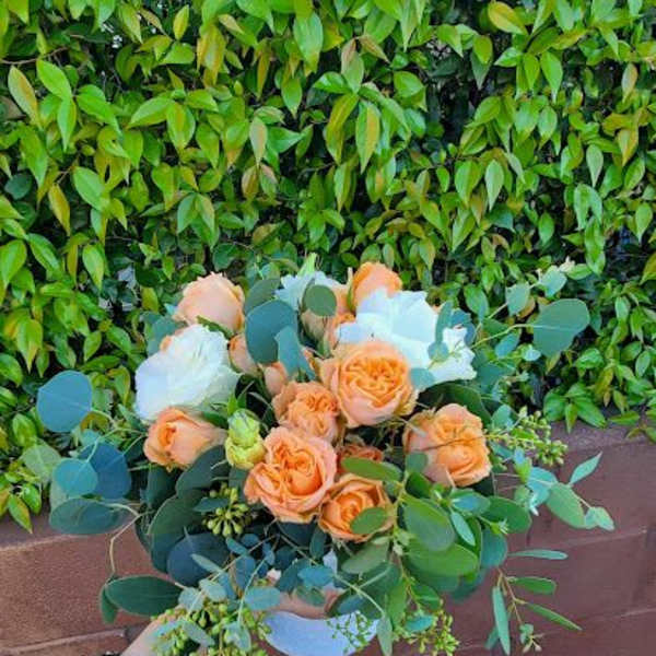 Low arrangement of peach roses and white blooms in a white bowl vase, held in front of a leafy hedge