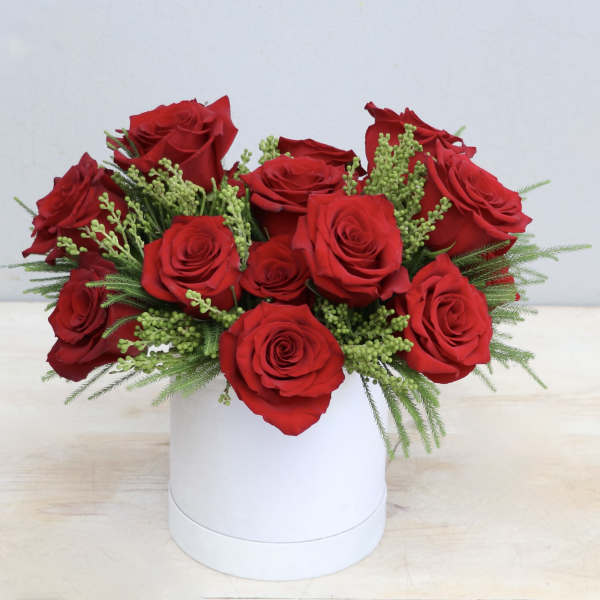 Low arrangement of red roses with green filler sprigs in a white round box.