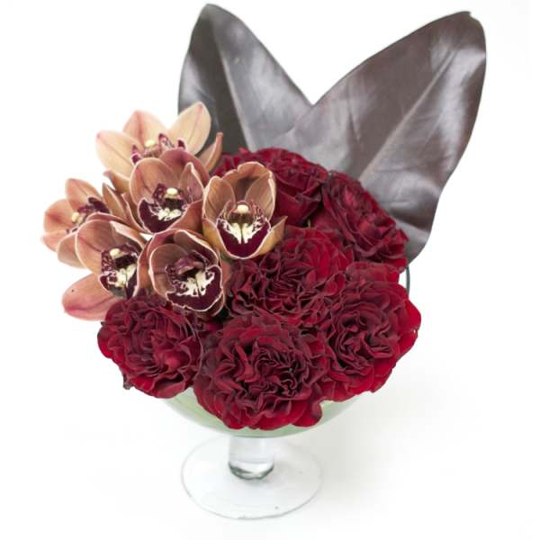 Red roses and orchids arranged in a glass vase with dark leaves.