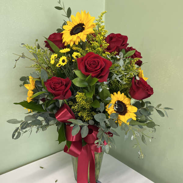 Tall vase of red roses and yellow sunflowers with small yellow daisies and a red bow.