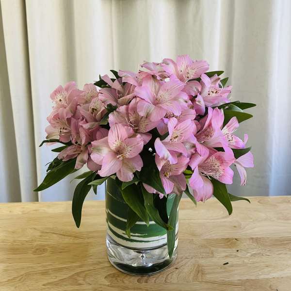 Compact bouquet of pink alstroemeria in a clear glass cylinder vase on a wooden table.