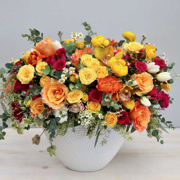 Low arrangement of yellow, orange, and red roses with ranunculus and tulips in a white ceramic bowl