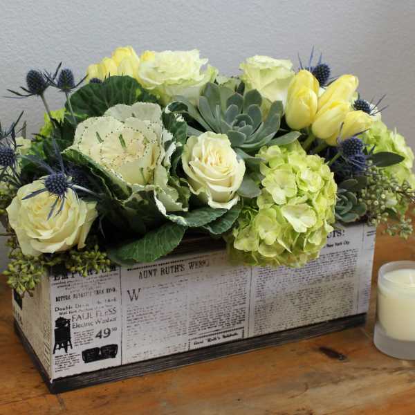 Rectangular floral arrangement with pale roses, hydrangea, succulents, and a candle in a printed box