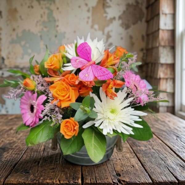 Bright arrangement of orange roses, pink blooms, and white mums in a metal bucket with a pink butterfly.