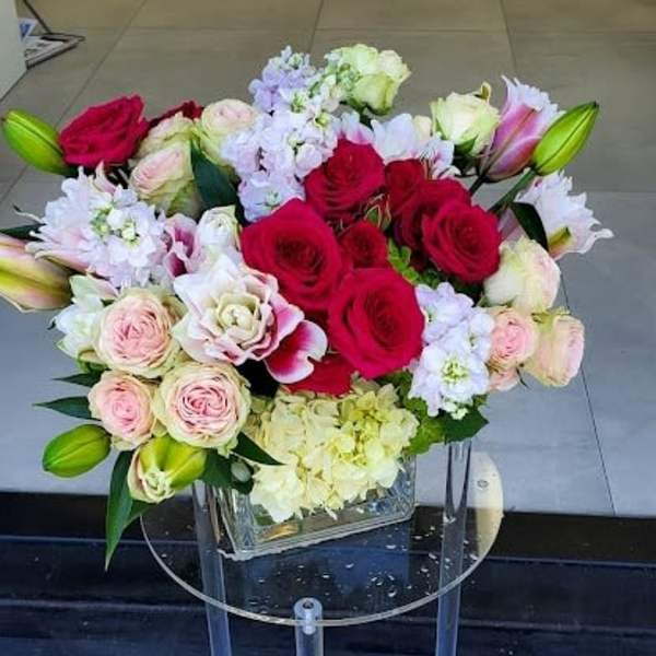 Low arrangement of red and pink roses, lilies, and light hydrangeas in a clear glass cube vase