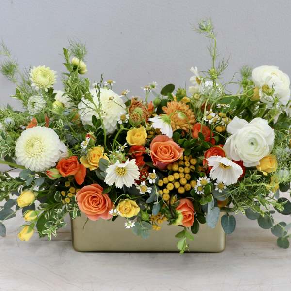 Low centerpiece of white dahlias, orange and yellow roses, and daisies in a gold rectangular vase