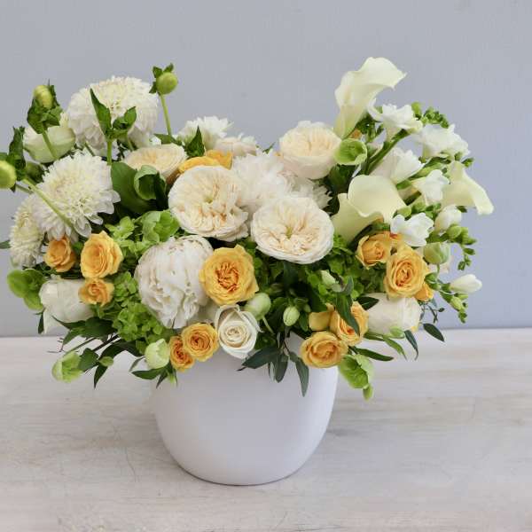 Low arrangement of white and yellow roses, dahlias, and calla lilies in a round white ceramic pot