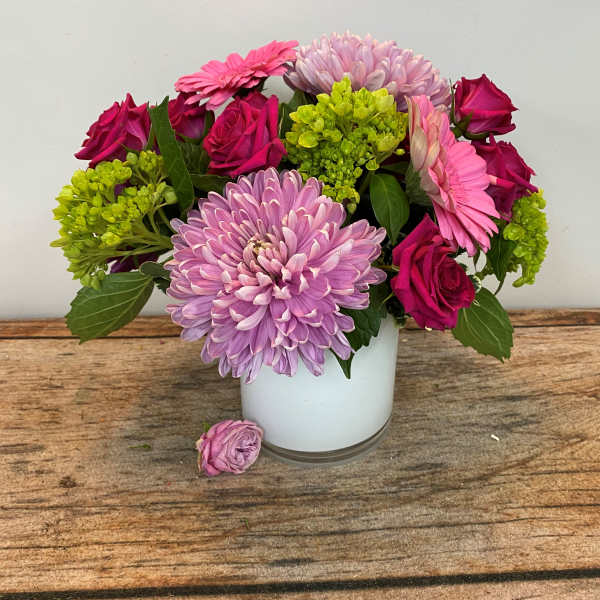 Pink roses, gerbera daisies, and lavender mum in a white vase on a wooden surface