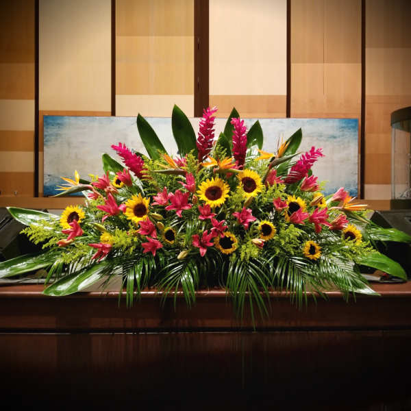 Large tropical arrangement with sunflowers, pink lilies, and tall pink blooms on a wooden surface