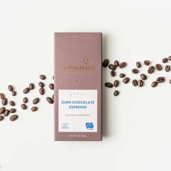Dark chocolate espresso bar in mauve wrapper with coffee beans scattered on a white surface