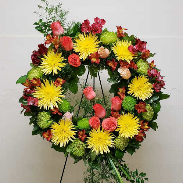 Standing wreath of yellow mums, pink roses, and red flowers on a green easel display