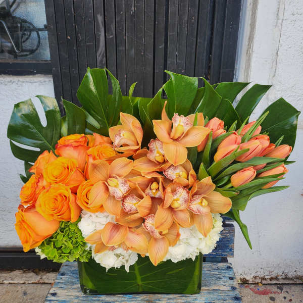 Orange roses and orchids arranged in a glass vase with large tropical leaves