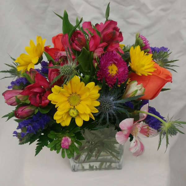 Mixed bouquet in a clear square vase with yellow daisies and pink, orange, and purple blooms