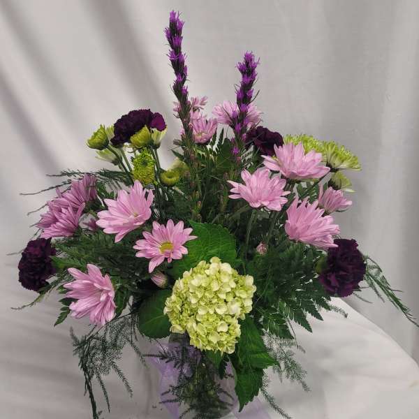 Pink daisies and purple carnations in a clear vase with a hydrangea bloom