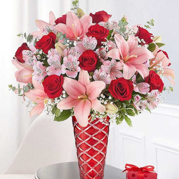 Pink lilies and red roses in a red vase with a small gift box