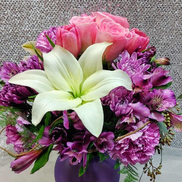 Pink roses and purple flowers arranged in a purple vase with a white lily