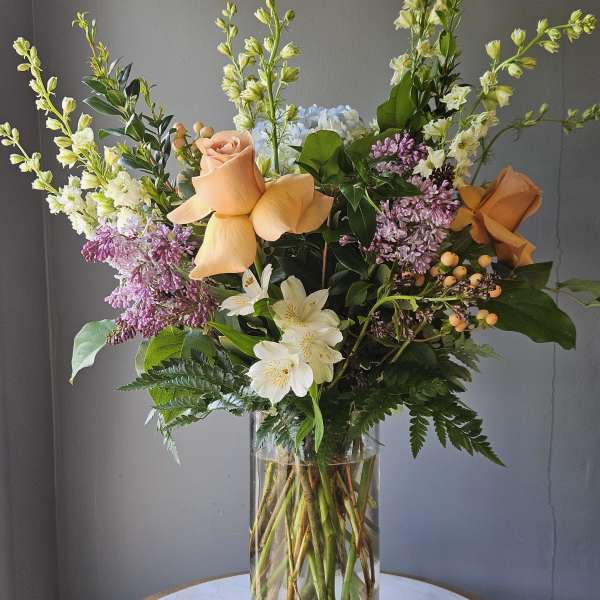Tall mixed bouquet with peach roses, white blooms, lilac stems in a clear glass cylinder vase.