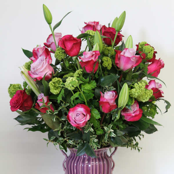 Tall arrangement of pink roses and lily buds in a mauve ceramic vase with green accent blooms