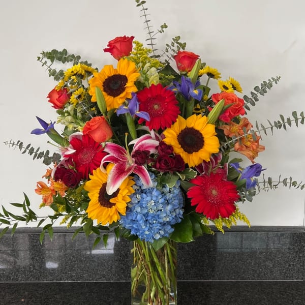 Mixed bouquet with sunflowers, roses, lilies, and blue hydrangea in a glass vase
