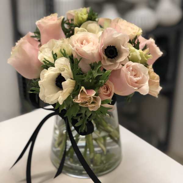 Blush pink roses and white anemones in a clear glass vase with a black ribbon bow.