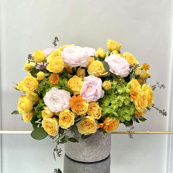 Yellow roses and soft pink blooms arranged with green hydrangeas in a round gray vase.