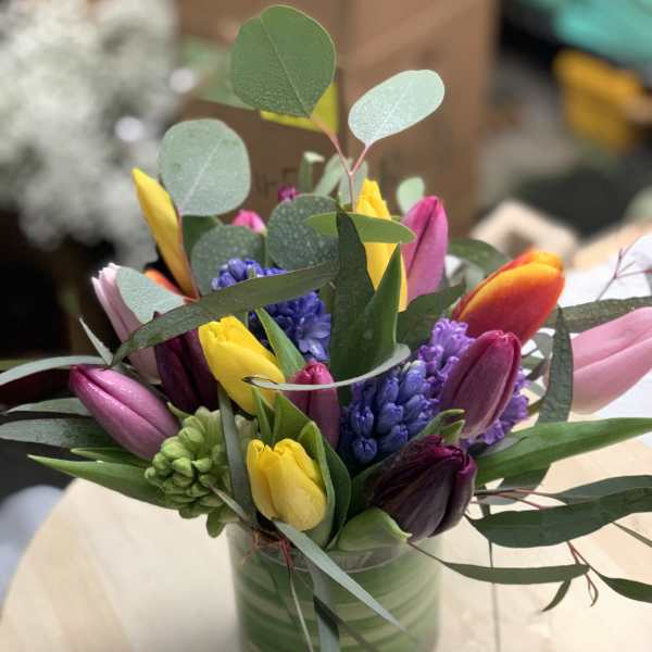Short glass vase filled with yellow, pink, and purple tulips with purple hyacinths on a light wood table