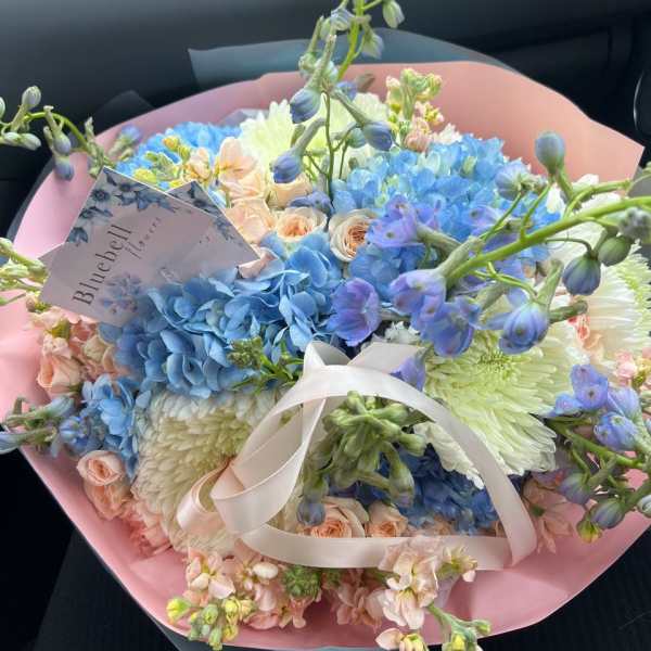 Pastel bouquet of blue hydrangeas, peach roses, and white blooms wrapped in pink paper
