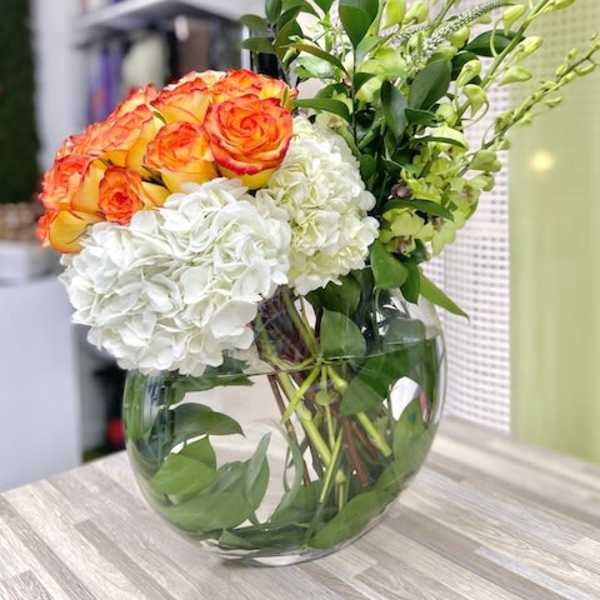 Round glass vase with orange roses, white hydrangeas, and tall green orchid stems