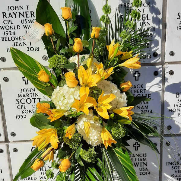 Tall spray of yellow lilies and roses with white hydrangeas on a stand in front of a marble memorial wall