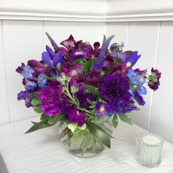 Round arrangement of purple and blue flowers in a clear glass vase on a white table.